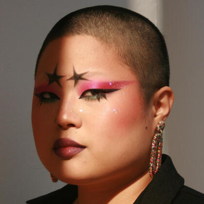 Josephine: a Korean queer person with a shaved head. Her makeup is sparkly and angular, making her largely unapproachable-looking. Half her face is cast in shadow. Her eyebrows are black, drawn-on stars.