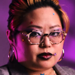 Headshot 2025 Josephine Josephine: a Korean queer person with a blonde hair in a purple lit room. She is glaring into the camera with purple eyeshadow and lipstick. She has star stickers on her face and gold hoop earrings.