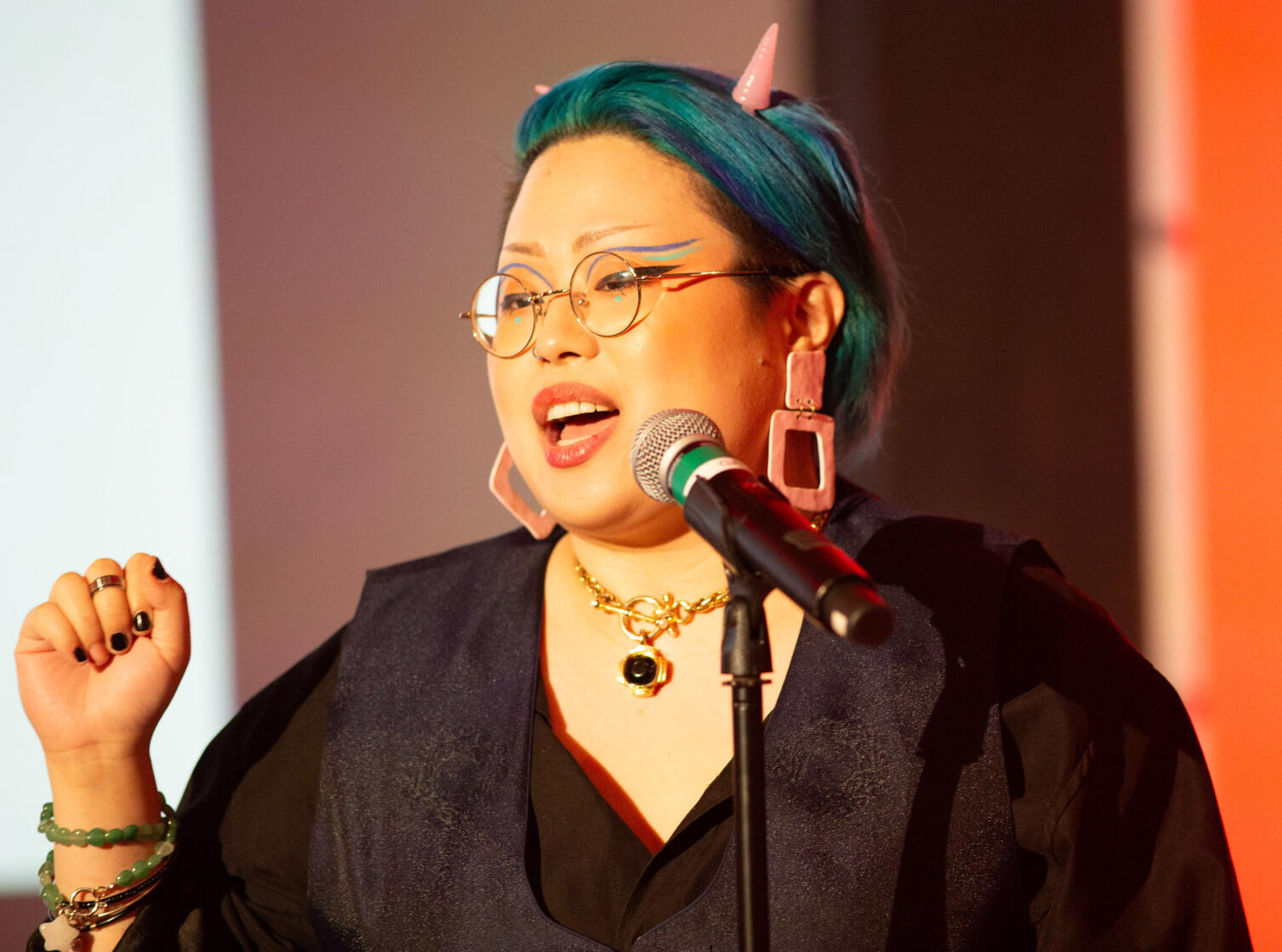 NJ Web Fest 2023 Josephine Josephine: a Korean queer person with blue hair, pink devil horn pins and big pink earrings. Her makeup is graphic blue eyeliner. She is at a microphone, mid-speaking.